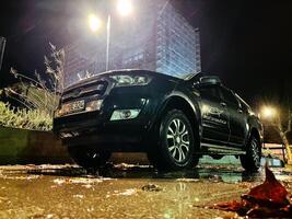 buda, montenegro - 25 diciembre 2022. negro suv soportes en el ligero de un linterna en mojado asfalto cerca un de muchos pisos edificio foto