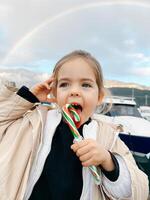 Little girl licks a lollipop against the backdrop of a rainbow over the mountains photo