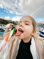 Little girl with her tongue hanging out reaches for a lollipop against the backdrop of a rainbow photo