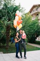 sonriente mamá abrazando papá con un pequeño niña en su brazos en el jardín cerca vistoso globos foto