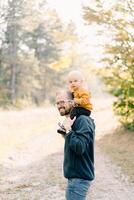 Smiling dad with a little boy on his shoulders stands half-turned in a clearing in the forest photo