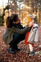 Mom puts a warm headband on little girl head while squatting in the autumn park photo