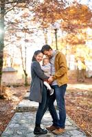 sonriente papá y mamá conmovedor frentes mientras en pie con un pequeño niña en su brazos en el otoño bosque foto