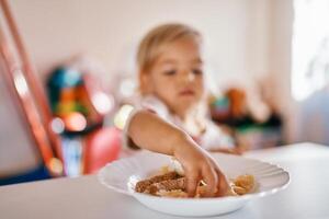 pequeño niña se sienta a el mesa y toma fuera pasta desde un plato con su manos foto