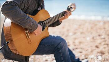 Musician plays the acoustic guitar while sitting on a chair on the beach. Cropped. Faceless photo