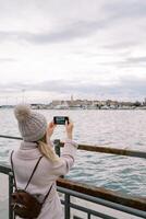 niña con un mochila soportes en el muelle a el barandilla y toma imágenes de casas en el costa con un teléfono inteligente espalda ver foto