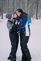 Mom and dad kiss a small child in their arms while standing in a snowy forest photo