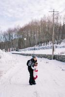 sonriente madre escoge arriba un pequeño niño en su brazos en un Nevado la carretera en el parque foto