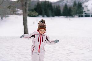 pequeño niña camina mediante un cubierto de nieve bosque, equilibrio su manos en mitones foto