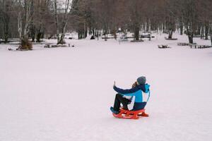 Dad taking smartphone selfie of himself and little kid sitting on sled on snowy lawn. Back view photo