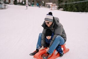 sonriente papá con un pequeño chico montando abajo un Nevado colina en un trineo foto