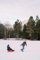 Dad is carrying mom with a child on a sled across a snowy plain looking back. Side view photo