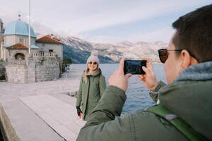 Man shoots woman with a smartphone on the pier near the Church of Our Lady of the Rocks. Back view. Montenegro photo