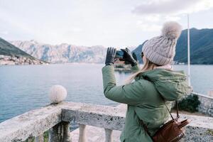 Girl with a backpack shoots the sea and a mountain range with a smartphone while standing at a stone balustrade. Side view photo