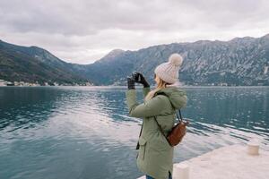 Woman stands on the pier and takes pictures of the sea and mountains. Side view photo