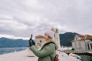 Woman stands on the island of Gospa od Skrpjela and takes pictures of the mountains with a smartphone. Side view. Montenegro photo