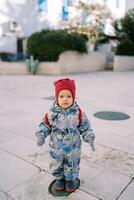 Little girl in overalls and a hat stands on a tile in the courtyard of the house photo