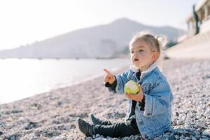 Little girl with an apple sits on a pebble beach and points to the sea photo