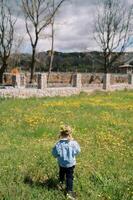 Little girl in a wreath stands on a flowering lawn and looks at a stone fence. Back view photo