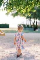 Little girl is walking on the playground under a green tree. Back view photo