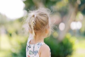 pequeño niña con fluido pelo soportes cerca un árbol y puntos a él. espalda ver foto