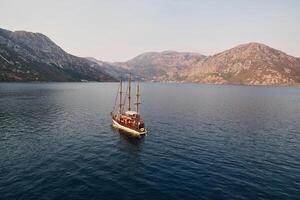 Wooden schooner sails across the sea towards a mountain range on the horizon. Drone photo