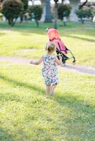 Little girl walks along a green lawn to a stroller standing on a path. Back view photo
