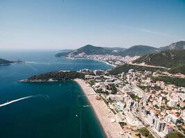 Sandy beach with sun umbrellas by the sea opposite high-rise hotels at the foot of the mountains. Budva, Montenegro. Drone photo