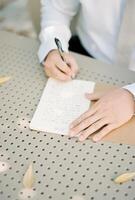 Groom writes an oath with a pen on a piece of paper while sitting at the table. Cropped. Faceless photo