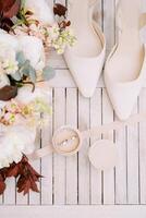 Wedding rings in a box stand on the table near the bride shoes and a bouquet of flowers. Top view photo