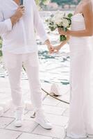 Groom with a microphone in his hand holds the hand of bride with a bouquet while standing on the pier. Cropped. Faceless photo