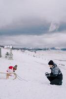 Mother and a little girl make snowballs while squatting near a sled in a clearing near the house photo