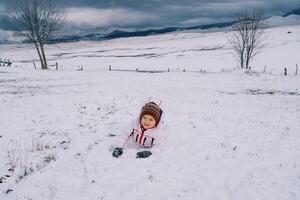 pequeño riendo niña mentiras en su estómago en el nieve en un montaña Valle foto