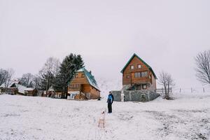 Dad stands near the house, looking at a small child climbing a hill with a sled photo