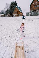 Little girl climbs a hill with her dad sitting on it with a sled on a rope, looking back. Back view photo