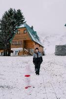 Small child walks towards his laughing mother along a snow-covered hill near a wooden cottage. Back view photo