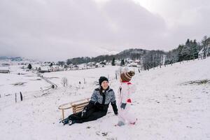 Little girl walks along a snowy hill towards her sitting smiling mother with a sleigh turned on its side photo