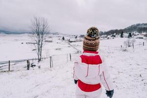 Little girl stands on a snowy hill and looks at the village. Back view photo