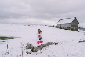 Little girl sits on a snowy pasture near a farm and looks away photo