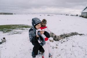 Mom hugs a little girl squatting on a snowy pasture near a farm photo