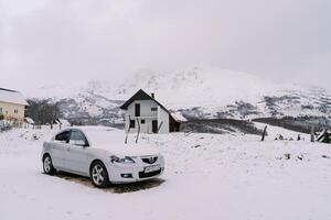 Snow-covered car with raised windshield wipers stands near a cottage in a mountain valley photo