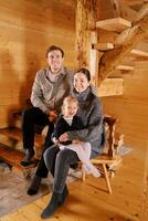 sonriente papá sentado en el escalera siguiente a mamá y pequeño niña sentado en un silla en un de madera cabaña foto