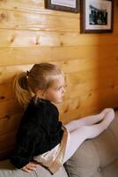 Little girl sits on the back of the sofa leaning on a wooden wall and looks into the distance photo