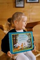 pequeño sonriente niña se sienta en un silla con un vistoso libro en su manos. título. cuentos en imágenes foto