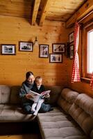 Mom reads a fairy tale to a little girl sitting in her arms on a corner sofa in a wooden chalet photo