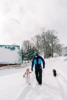 negro perro sigue un papá caminando mano en mano con un pequeño niña a lo largo un Nevado la carretera en el pueblo foto