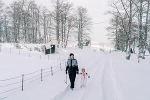 madre y un pequeño niña caminar participación manos a lo largo un Nevado la carretera en un pueblo en el borde de un bosque foto