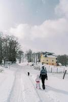 Mother with a small child walks holding hands through a snowy village behind a black dog. Back view photo