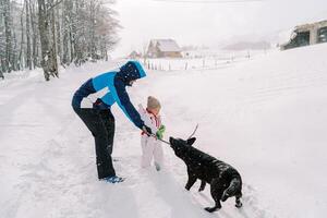 papá obras de teatro con un negro perro con un palo mientras en pie con un pequeño niña en un Nevado pueblo la carretera foto