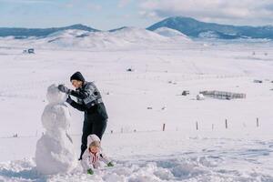 Little girl picks in the snow, bending over next to her mother making a snowman photo